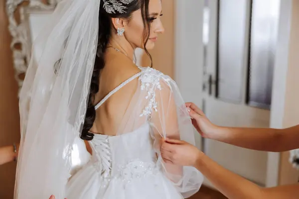 The bride's friend helps her get dressed in a white wedding dress in the morning. wedding morning, blurred focus. A friend of the bride helps to tie her wedding dress.