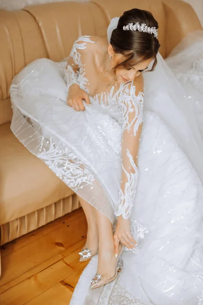 Young bride in beautiful wedding dress putting on shoes indoors. Bride dresses shoes before the wedding ceremony. Detail of bride putting on high heeled sandal wedding shoes. Wedding bride shoes.