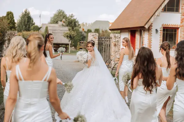 Wedding photography. A brunette bride in a long wedding dress and her friends in beige dresses walk to the houses, rear view. Autumn wedding. Young girls. Posing
