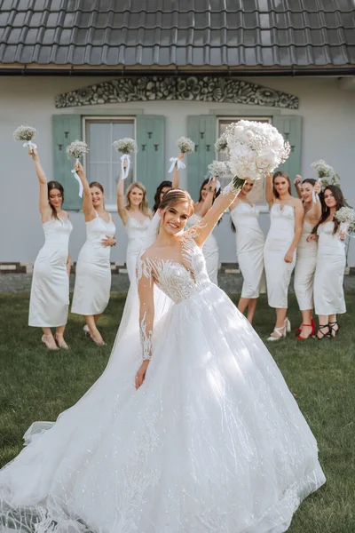 Wedding photography. The bride in a long wedding dress with a veil, holding a bouquet, poses in front of her friends in white dresses against the background of the building. Friendship. A group of young girls. celebration.