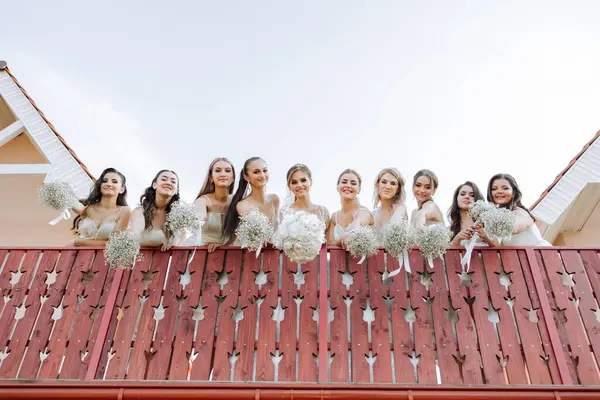 Wedding photo session in nature. The bride and her bridesmaids pose with bouquets on a balcony with a red railing. Photo from below. Friendship. A group of young girls. celebration.