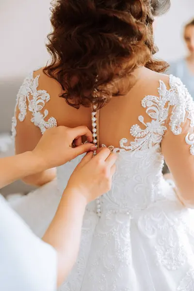 The bride\'s friend helps her get dressed in a white wedding dress in the morning. wedding morning, blurred focus. A friend of the bride helps to tie her wedding dress.