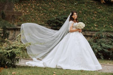 Şehvetli beyaz elbiseli esmer gelin elinde buket, poz veriyor. Duvak havaya fırlatıldı. Güzel saç ve makyaj. Doğada düğün fotoğrafı çekimi.