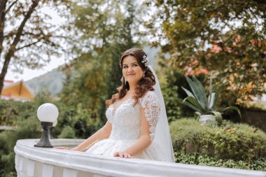 Düğün portresi. Uzun kollu, dantel elbiseli esmer gelin, korkulukların yanında poz veriyor. Güzel saç ve makyaj. Sonbahar. Gün ışığı. kutlama.