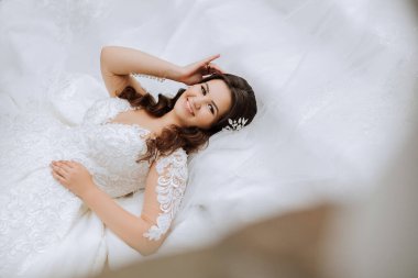 Esmer bir gelin dantelli bir elbise ve tacın üzerine uzanmış saçlarına dokunuyor. Güzel makyaj ve saç. Beyaz bir arka planda. Güneşli bir gün. Düğün töreni.