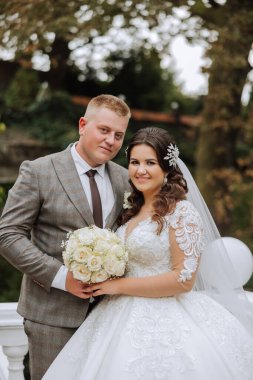 Şık, genç bir damat ve uzun beyaz bir elbise içinde güzel bir gelin ve ellerinde bir buket ile uzun bir duvak, sonbahar doğasında parkta sarılıyorlar. Yeni evlilerin düğün portresi..