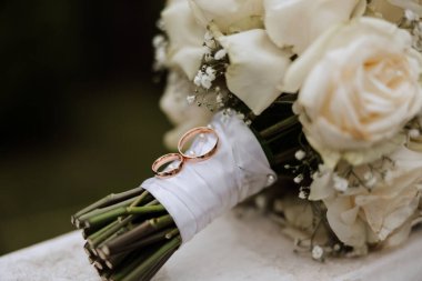 Bir buket beyaz gül doğada duruyor. Arka planı bulanık ve alyansları da yanında..