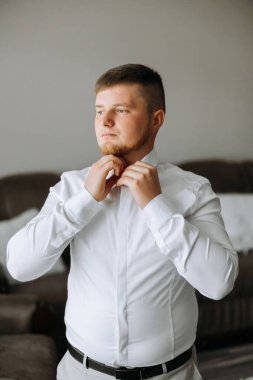 Beyaz gömlekli bir adam odanın penceresinin yanında durur ve yakasındaki ve kollarındaki düğmeleri bağlar. Hazır ol. Bir adamın şık bir iş portresi, yakın çekim fotoğrafı. Damat hazırlanıyor..