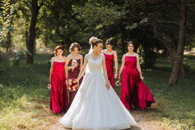 Düğün fotoğrafçılığı. Beyaz elbiseli, buketli esmer bir gelin ve kırmızı elbiseli esmer arkadaşları bahçede kırmızı buketlerle poz veriyorlar. Arkadaşlık. Bir grup genç kız. Kutlama.