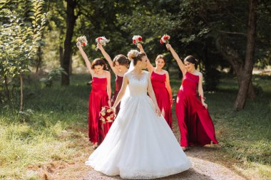 Düğün fotoğrafçılığı. Çiçekli, beyaz elbiseli esmer bir gelin ve kırmızı elbiseli esmer arkadaşları ellerinde kırmızı buketlerle bahçede poz veriyorlar. Arkadaşlık. Bir grup genç kız. Kutlama.