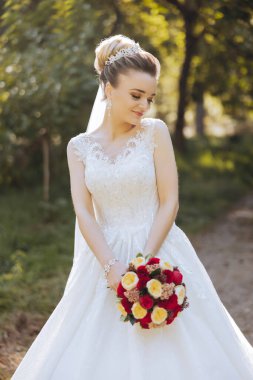 Düğün fotoğrafı. Dantelli, dolgun elbiseli esmer bir gelin elinde bir buket tutarak bahçede yürüyor, içtenlikle gülümsüyor. Harika saç ve makyaj. Moda ve tarz. Kutlama.