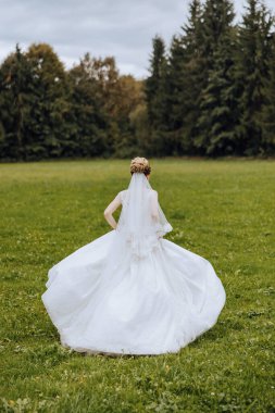 Beyaz elbiseli, uzun trenli esmer gelin elinde buket, bahçede yürüyor. Doğada düğün fotoğrafı çekimi. Güzel saç ve makyaj. Kutlama