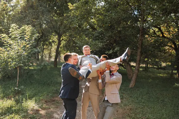 Wedding photo session in nature. Friends hold the groom in their arms, have fun, pose in the forest. Sincere smile. A group of young men in suits. Style. Friendship