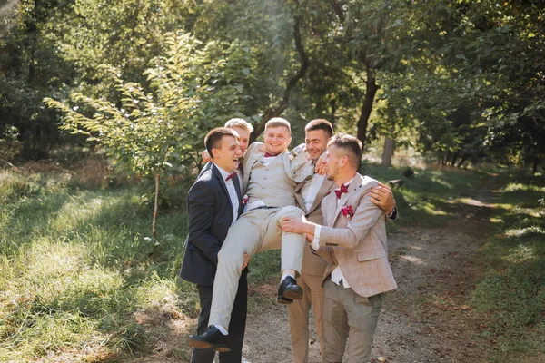 Wedding photo session in nature. Friends hold the groom in their arms, have fun, pose in the forest. Sincere smile. A group of young men in suits. Style. Friendship