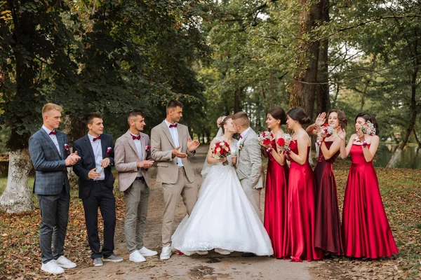 Wedding photo session in nature. The bride and groom kiss and their friends applaud them, posing against the background of trees. Happiness. A group of young people. Celebration. Autumn wedding