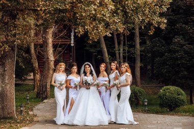 Düğün fotoğrafçılığı. Beyaz elbiseli, buketli esmer bir gelin ve beyaz elbiseli esmer arkadaşları ormanda poz veriyor. Sonbahar düğünü. Arkadaşlık. Bir grup genç kız. Kutlama.