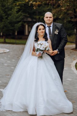 Şık, genç bir damat ve uzun beyaz bir elbise içinde güzel bir gelin ve ellerinde bir buket ile uzun bir duvak, sonbahar doğasında parkta sarılıyorlar. Yeni evlilerin düğün portresi..