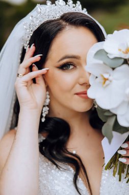 Portre. Elbise ve duvak takmış esmer bir gelin şık bir taç poz verir ve buketini koklar. Gümüş mücevherler. Güzel makyaj ve saç. Sonbahar düğünü. kutlama