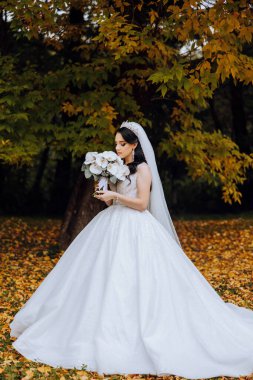 Beyaz elbiseli, uzun trenli esmer gelin elinde buket, bahçede yürüyor. Doğada düğün fotoğrafı çekimi. Güzel saç ve makyaj. Kutlama