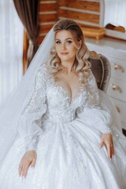 Şık bir gelinlik içinde sarı saçlı ve düğün sabahı odasında çarpıcı makyajlı güzel bir gelinin moda fotoğrafı. Gelin düğün için hazırlanıyor.