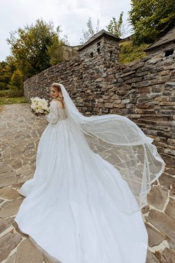 Şehvetli beyaz elbiseli sarışın gelin elinde buket, poz veriyor. Duvak havaya fırlatıldı. Güzel saç ve makyaj. Doğada düğün fotoğrafı çekimi. Elbisenin uzun treni.