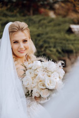 Düğün portresi. Dantel elbiseli kıvırcık sarışın gelin, elinde buketle dışarıda poz veriyor. Güzel saç ve makyaj. Gün ışığı. Kutlama