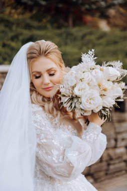 Düğün portresi. Dantel elbiseli kıvırcık sarışın gelin, elinde buketle dışarıda poz veriyor. Güzel saç ve makyaj. Gün ışığı. Kutlama