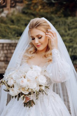 Düğün portresi. Dantel elbiseli kıvırcık sarışın gelin, elinde buketle dışarıda poz veriyor. Güzel saç ve makyaj. Gün ışığı. Kutlama