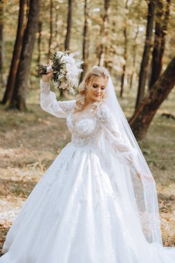 Düğün portresi. Sarışın gelin bir buket tutuyor ve doğada poz veriyor. Havaya uzun bir duvak atıldı. Güzel eller. Dantelli elbise.