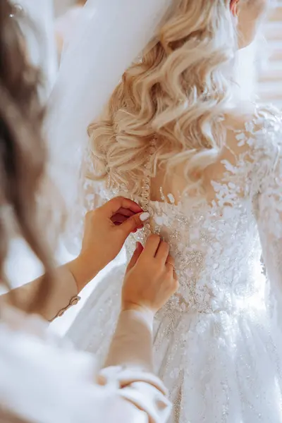 The bride\'s friend helps her get dressed in a white wedding dress in the morning. wedding morning, blurred focus. A friend of the bride helps to tie her wedding dress.