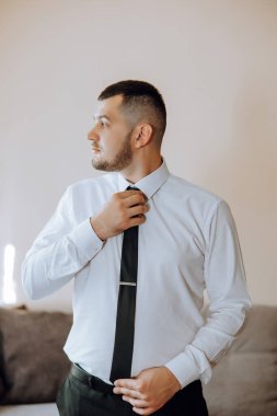 Beyaz gömlekli genç bir adam odanın penceresinin yanında duruyor ve kravat takıyor. Damat sabah giyinir ve düğün için hazırlanır..