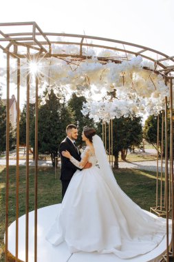 Takım elbiseli şık bir damat ve beyaz dantel elbiseli şirin bir gelin sonbahar doğasının ve güneş ışınlarının arka planına karşı çiçeklerden yapılmış bir kemerde dururlar ve birbirlerine bakarlar..