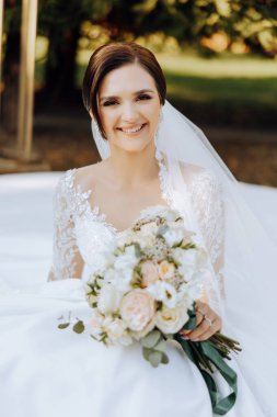 Esmer gelin, tüm samimiyetiyle gülümseyerek yerde açık elbisesiyle oturuyor ve elinde bir buket çiçek tutuyor. Düğün töreni. Güneşli bir gün. Güzel saç ve makyaj.