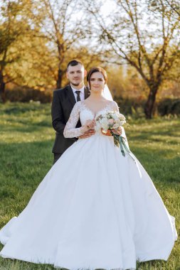 Şık, genç bir damat ve uzun beyaz bir elbise içinde güzel bir gelin ve ellerinde bir buket ile uzun bir duvak, sonbahar doğasında parkta sarılıyorlar. Yeni evlilerin düğün portresi..