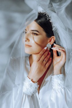 Evdeki düğün töreninden önce güzel genç gelin küpelerini takar. Sabahları beyaz cüppeli bir gelin odasına küpe takıyor..