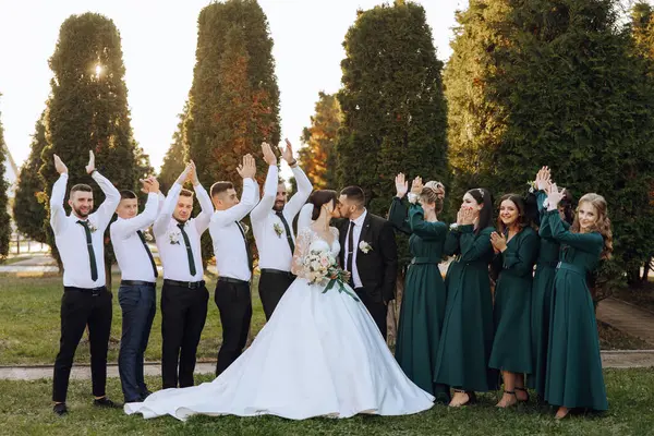 Wedding photo session in nature. The bride and groom kiss and their friends applaud them, posing against the background of trees. Happiness. A group of young people. Celebration. Autumn wedding