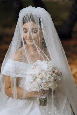 Düğün portresi. Esmer bir gelin elinde bir buket tutuyor ve uzun bir peçeyle doğada poz veriyor. Güzel makyaj. Dantelli elbise.