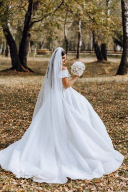 Beyaz elbiseli, uzun trenli esmer gelin elinde buket, bahçede yürüyor. Doğada düğün fotoğrafı çekimi. Güzel saç ve makyaj. Kutlama