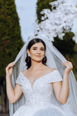 Düğün portresi. Esmer bir gelin, uzun bir peçeyle örtünerek doğada poz veriyor. Güzel makyaj. Dantelli elbise. Sonbahar fotoğraf çekimi