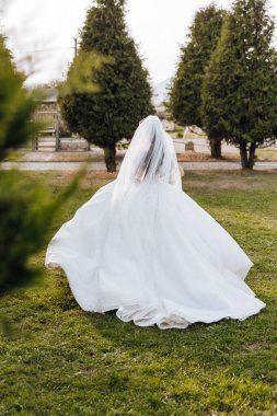 Beyaz elbiseli, uzun trenli esmer gelin elinde buket, bahçede yürüyor. Doğada düğün fotoğrafı çekimi. Güzel saç ve makyaj. Kutlama