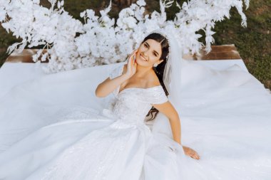 Duvak takmış esmer bir gelin ve saçında bir taç geniş bir elbisenin üzerine oturmuş, daire şeklinde çiçeklerden yapılmış beyaz bir kemerin yanında poz veriyor. Beyaz bir arka planda. Güneşli bir gün. Düğün töreni.