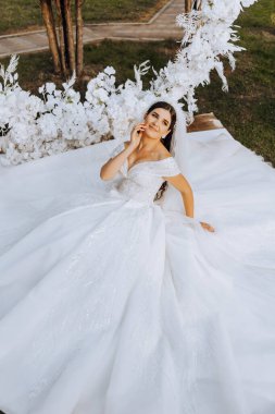 Duvak takmış esmer bir gelin ve saçında bir taç geniş bir elbisenin üzerine oturmuş, daire şeklinde çiçeklerden yapılmış beyaz bir kemerin yanında poz veriyor. Beyaz bir arka planda. Güneşli bir gün. Düğün töreni.