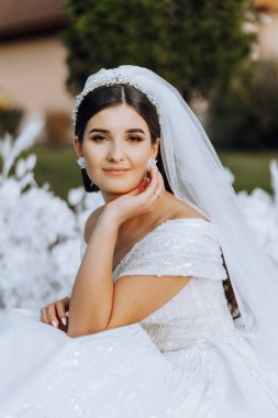 Esmer gelin, tüm samimiyetiyle gülümseyerek, çiçeklerden yapılmış beyaz bir kemerin yanında, katlanmış bir elbiseyle yerde oturuyor. Düğün töreni. Güneşli bir gün. Güzel saç ve makyaj.