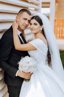 Şık, genç bir damat ve uzun beyaz bir elbise içinde güzel bir gelin ve ellerinde bir buket ile uzun bir duvak, sonbahar doğasında parkta sarılıyorlar. Yeni evlilerin düğün portresi..