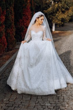 Uzun trenli beyaz elbiseli esmer bir gelin elbiseyi tutar ve taş patikadan aşağı doğru yürür. Sonbahar. Doğada düğün fotoğrafı çekimi. Güzel saç ve makyaj. kutlama