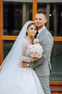 Şık, genç bir damat ve uzun beyaz bir elbise içinde güzel bir gelin ve ellerinde bir buket ile uzun bir duvak, sonbahar doğasında parkta sarılıyorlar. Yeni evlilerin düğün portresi..