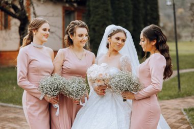 Düğün fotoğrafçılığı. Beyaz elbiseli, buketli esmer bir gelin ve pembe elbiseli esmer arkadaşları bahçede çingene buketleriyle poz veriyorlar. Arkadaşlık. Bir grup genç kız. Kutlama.