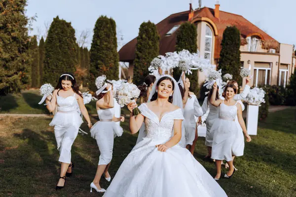 Wedding photography. The bride in a wedding dress and her friends in white dresses pose with bouquets near a white arch. Wedding ceremony. The same clothes. Friendship. A group of young girls. Celebration.