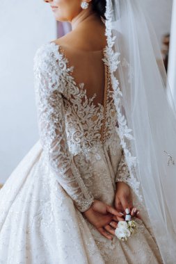 Genç gelin arka tarafta duruyor. Gelinlik giymiş güzel bir kadın, pencerenin yanındaki bir odada. Uzun trenli beyaz gelinlikli çekici kız.