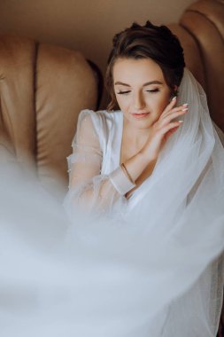 Yatak odasında beyaz bir bornozla inanılmaz güzel bir kızın portresi. Gelin peçesini elinde tutuyor ve kendini onunla örtüyor..